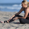 Woman sitting on the beach wearing black leggings with a visible brand logo.